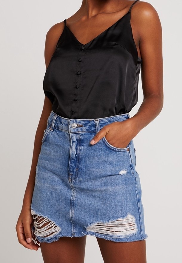 Femme portant un caraco noir en satin boutonné et une mini jupe en jean bleu délavé, la main dans la poche.