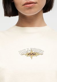 Κοντινό πλάνο ενός ατόμου που φοράει πουκάμισο εκρού με κίτρινο ήλιο και το λογότυπο "BILLABONG" τυπωμένο στο στήθος.