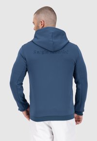 Felpa con cappuccio blu, caratterizzata da pannelli delle maniche texturizzati, logo in rilievo sulla schiena e design aderente. Tessuto liscio con polsini elasticizzati.