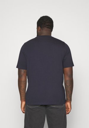 Homme aux cheveux courts et bouclés avec des tatouages aux bras, portant un t-shirt bleu marine uni à manches courtes et un pantalon gris foncé, vu de dos.