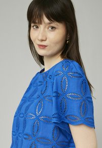 Haut bleu brodé avec découpes circulaires et manches festonnées. Fabriqué en tissu léger avec une finition texturée.