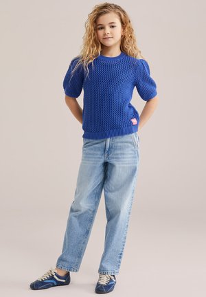 Junges Mädchen mit lockigem blondem Haar steht mit verschränkten Händen hinter dem Rücken, trägt ein blaues, gestricktes Kurzarmoberteil, hellblaue Jeans und marineblaue Turnschuhe.
