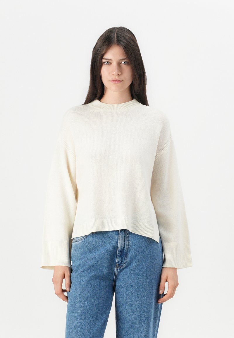 Maglione a maglia color crema con scollatura tonda, spalle scese e maniche larghe. L'orlo è accorciato sopra la vita. Abbinato a jeans blu.