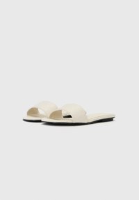Sandales plates à bout ouvert, fabriquées en matériau texturé crème, avec une semelle noire et une esthétique minimaliste.