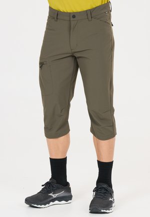 Mann trägt olivgrüne Dreiviertelhose mit Reißverschlusstasche, schwarze Socken und schwarze Laufschuhe.