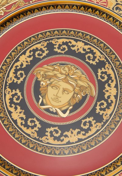 Κεραμικό πιάτο με κόκκινο και μαύρο σχέδιο. Κεντρική χρυσή ανάγλυφη απεικόνιση προσώπου με ρέοντα μαλλιά, περιβαλλόμενη από περίπλοκα χρυσά και μαύρα μοτίβα.