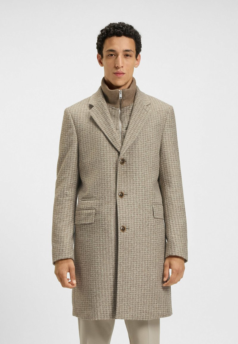 Cappotto beige a motivo houndstooth con vestibilità su misura, rever a punta, due tasche frontali e bottoni marroni, indossato sopra un maglione a collo alto in knit.