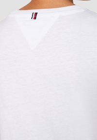 Biała bawełniana koszulka z dekoltem w kształcie litery V z tyłu, z małą czarną, czerwoną i białą paskowaną metką. Gładka tekstura.