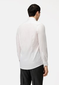 Chemise blanche à manches longues en tissu léger, avec un col classique, un design de dos empièce et des poignets boutonnés avec trois boutons.