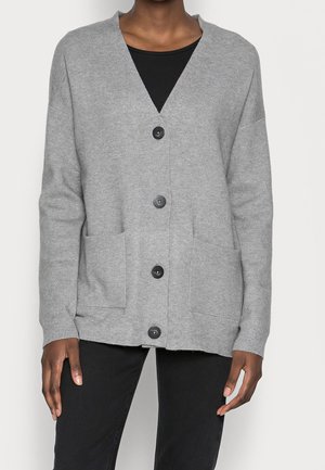 Osoba ubrana w jasnoszary zapinany na guziki kardigan z dwoma przednimi kieszeniami, na czarnej bluzce i czarnych spodniach, stojąca na jednolitym tle.