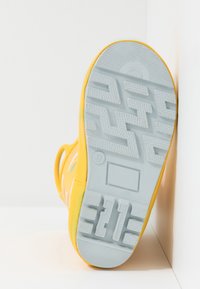 Żółte trampki z gumową podeszwą; teksturowana szara podeszwa z geometrycznym wzorem. Minimalistyczny design, bez widocznych sznurowadeł, skoncentrowany na trwałości.