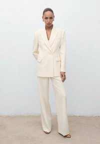 Crème gekleed pak met een lange blazer met één knoop, wijde broekspijpen en een gestructureerde stof. Gouden oorbellen als accenten.