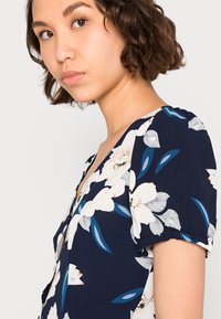 Vestido floral com fundo azul-marinho e flores brancas e azuis. Feito de um tecido macio, com mangas curtas bufantes e um decote em V profundo.