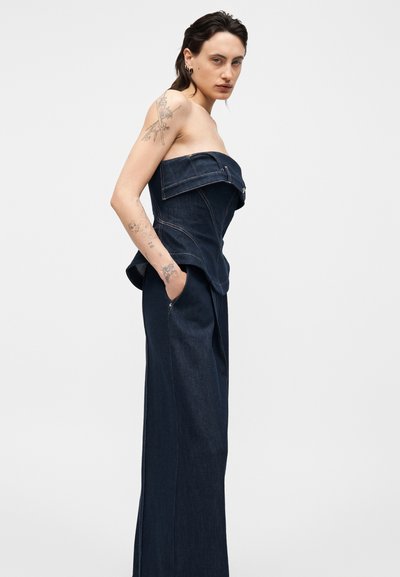 Tetoválásokkal rendelkező nő vállpánt nélküli sötét farmer fűzőt és hozzá illő, magas derekú, bő szárú nadrágot visel, kezét a zsebében tartva pózol egy egyszerű háttér előtt.