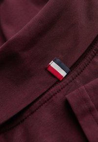 Burgundy tyg med ribbad textur, med en liten etikett i rött, vitt och blått. Sömmen är snygg och jämn.