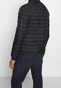 Man i en svart vadderad jacka och mörkblå jeans, som står med ryggen mot en enfärgad vit bakgrund.