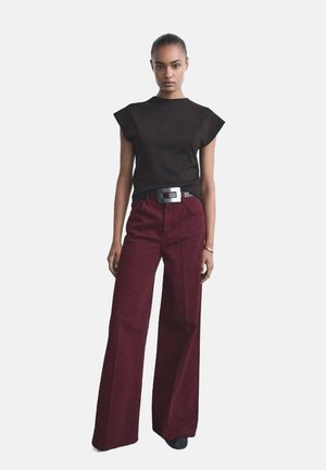 Top negro de manga corta con volantes en los hombros, combinado con pantalones burdeos de tiro alto y perneras anchas. La modelo lleva un cinturón con hebilla plateada.