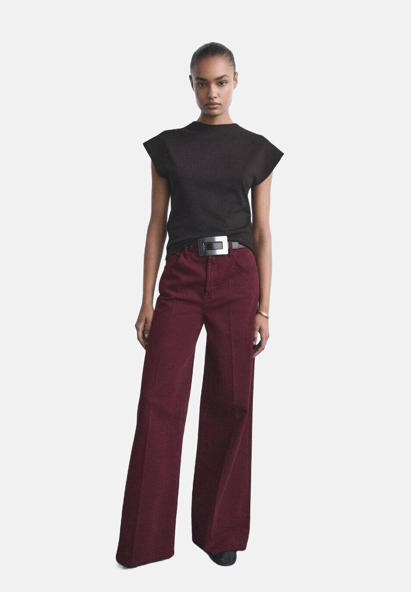 Haut noir à manches courtes avec des volants aux épaules, associé à un pantalon bordeaux à taille haute et à jambes larges. Le modèle porte une ceinture avec boucle en argent.