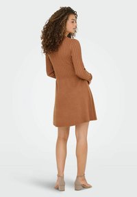 Pletena obleka v topli rjavi barvi z rebrastim vzorcem na vrhu in razširjenim krilom. Ima dolge rokave in prikrajšano pas, oblečena z visokimi sandalsi.