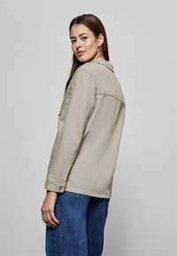 Beige denimjacka med krage, knäppte ärmslut och en enda bröstficka, i kombination med blå vida jeans. Slät struktur, avslappnad design.