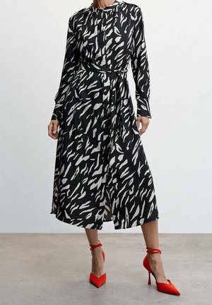 Schwarz-weiß gemustertes Kleid mit langen Ärmeln, tailliertem Bund und fließendem Rock; kombiniert mit roten spitz zulaufenden Absätzen.