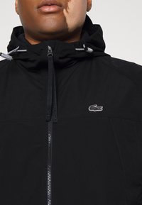 Svart zip-up jacka med huva, med en texturerad tyg, silverdetaljer och en liten Lacoste-logotyp på bröstet.