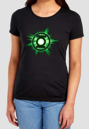 Frau trägt schwarzes T-Shirt mit leuchtend grünem Green Lantern-Logo und hellblauen Jeans, steht vor einfarbigem Hintergrund.