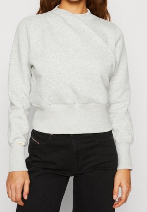 Femme portant un sweat court à manches longues gris clair et un jean noir taille haute, debout devant un fond uni.