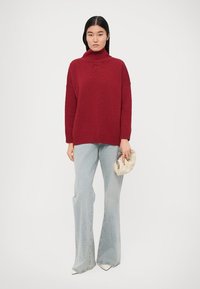 Pull à col roulé tricoté rouge avec une coupe ample, associé à un jean flare bleu clair et un sac clutch crème.