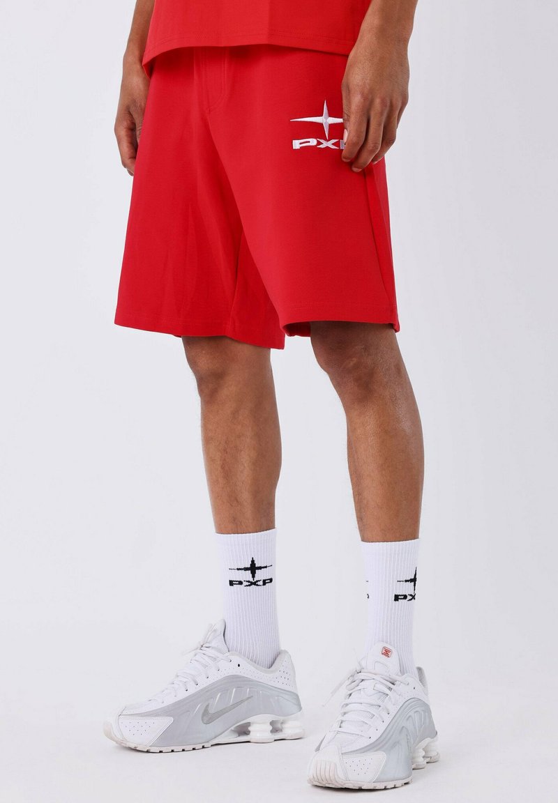 Homme portant un short rouge avec le logo PXG, des chaussettes crew blanches marquées PXG, et des baskets Nike blanches, se tenant devant un fond blanc.