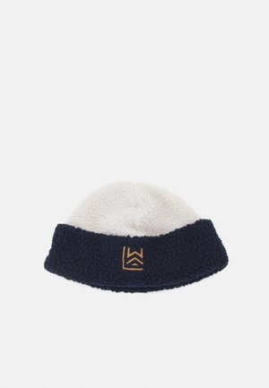Bonnet en polaire avec un dessus crème et un revers en marine foncé. Il présente une surface texturée et un petit logo doré sur le revers.