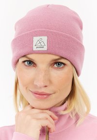 Gorro de punto acanalado rosa con una etiqueta de tela blanca que presenta un diseño de montaña y el texto "protesta". Textura suave, diseño ajustado.