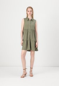 Robe sans manches vert olive avec un col, patte de boutonnage à l'avant et jupe à volants. Portée avec des talons strap de couleur beige. Design simple et décontracté.