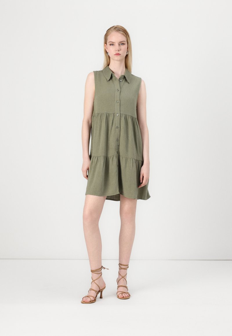Robe sans manches vert olive avec un col, patte de boutonnage à l'avant et jupe à volants. Portée avec des talons strap de couleur beige. Design simple et décontracté.