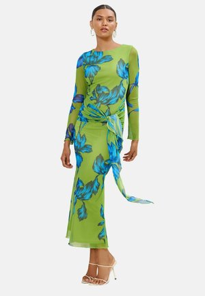 Frau trägt ein langes grünes Kleid mit blauem Blumenmuster und langen Ärmeln, gebundene Details an der Taille und weiße Sandalen mit hohen Absätzen.