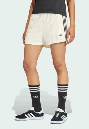 Osoba nosząca białe spodenki Adidas z czarnymi paskami, czarne skarpetki Adidas z białymi paskami oraz czarno-białe buty sportowe Adidas.