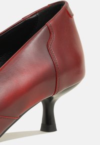 Zapato de tacón alto de cuero rojo con punta redondeada, detalle de costura curvada y un tacón negro delgado. Textura suave con un acabado pulido.