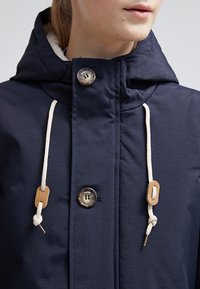 Giacca blu navy con cappuccio, caratterizzata da chiusura con bottoni, coulisse crema e dettagli in legno. Tessuto dalla texture liscia e vestibilità sartoriale.