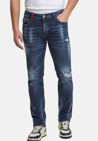 Hombre que lleva puestos unos jeans ajustados desgastados de color azul oscuro con salpicaduras de pintura blanca, combinados con zapatillas negras y blancas y una camisa blanca.