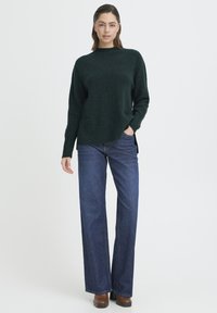 Pulover verde închis tricotat, cu gât înalt și tăieturi pe laterale, asortat cu blugi din denim albastru cu picioare largi și cizme maro până la gleznă.