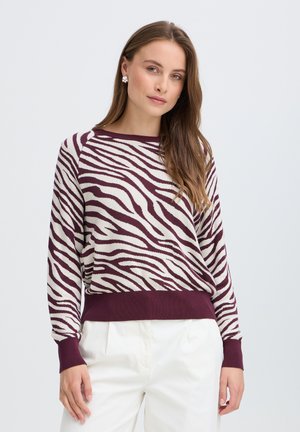 Femme aux longs cheveux bruns porte un pull zébré bordeaux et blanc et un pantalon blanc, debout devant un fond clair uni.