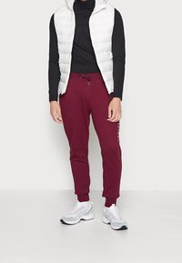 Man i vit pufferväst, svart långärmad tröja, vinröda joggers och vita sneakers som står mot en enkel bakgrund.