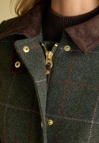 Giacca verde in misto lana con collo in suede marrone, zip dorata, bottoni a pressione e motivo a quadri con linee maroon e lilla chiaro.