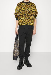 Sudadera negra con un patrón amarillo en espiral, pantalones negros y botas negras; sosteniendo una bolsa tote con un diseño oscuro y acentos de texto.