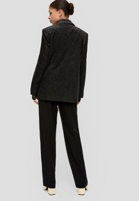Vrouw met haar in een bun, in een donkere, gestructureerde blazer, zwarte wijde broek en crèmewitte blokhak schoenen, met de rug naar de camera gekeerd tegen een effen achtergrond.