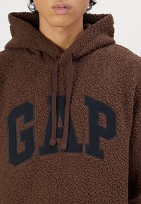 Hnědá fleecová mikina s kapucí, vybavená přední kapsou a černým logem "GAP". Látka má texturovaný, měkký vzhled a kapuci se šňůrkou na stažení.