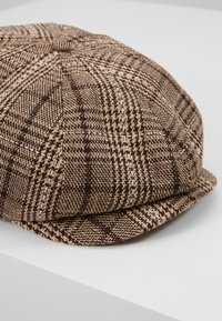 Casquette plate à carreaux marron et beige avec un détail bouton sur le dessus, posée sur une surface blanche.