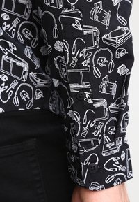 Camisa de tela negra con estampado gráfico blanco de dispositivos electrónicos; cuenta con un puño abotonado y bocetos detallados de gadgets y conectores.