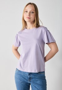 T-shirt di cotone lilla con maniche corte a sbuffo, scollatura rotonda e vestibilità rilassata. Abbinato a jeans in denim blu. Texture liscia, design casual.