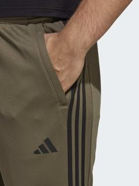 Olīvu zaļas sporta bikses ar melnām sānu svītrām un Adidas logotipu. Biksēm ir kabata gurnā, tās izgatavotas no gludas, vieglas auduma.